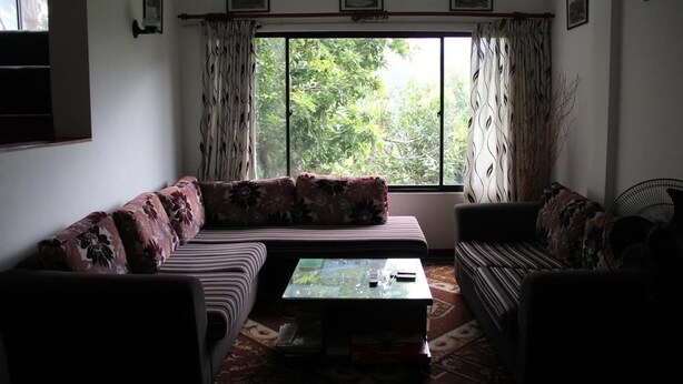 A living room with a sofa, a coffee table, and a fan. There are curtains on the windows, and a picture frame is visible on the wall. - Nature Villa 107 Kandy - Hostel (Kandy)