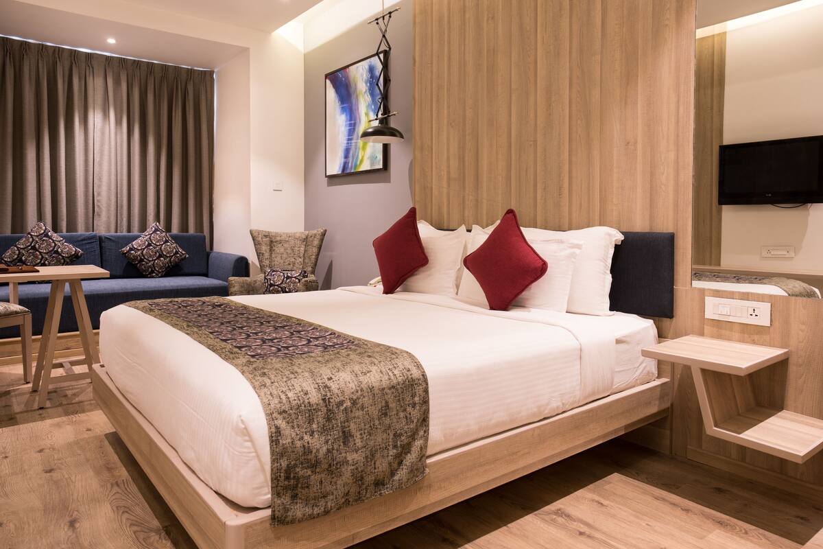 Moderni hotellihuone, jossa on suuri sänky, sohva, pieni pöytä ja seinälle asennettu televisio.