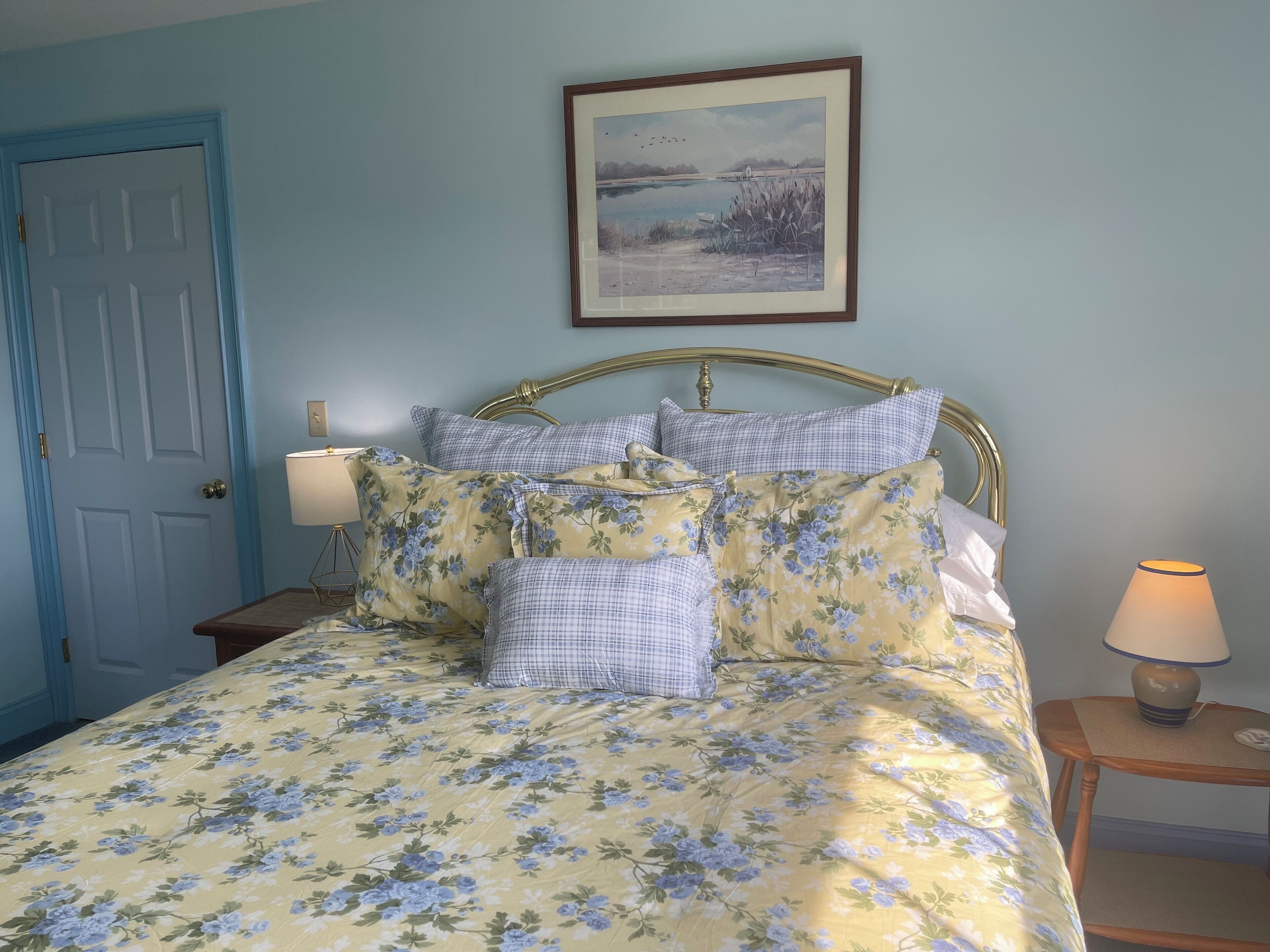 A bedroom with a floral bedspread, a checkered pillow set, a framed landscape painting, and a bedside lamp.