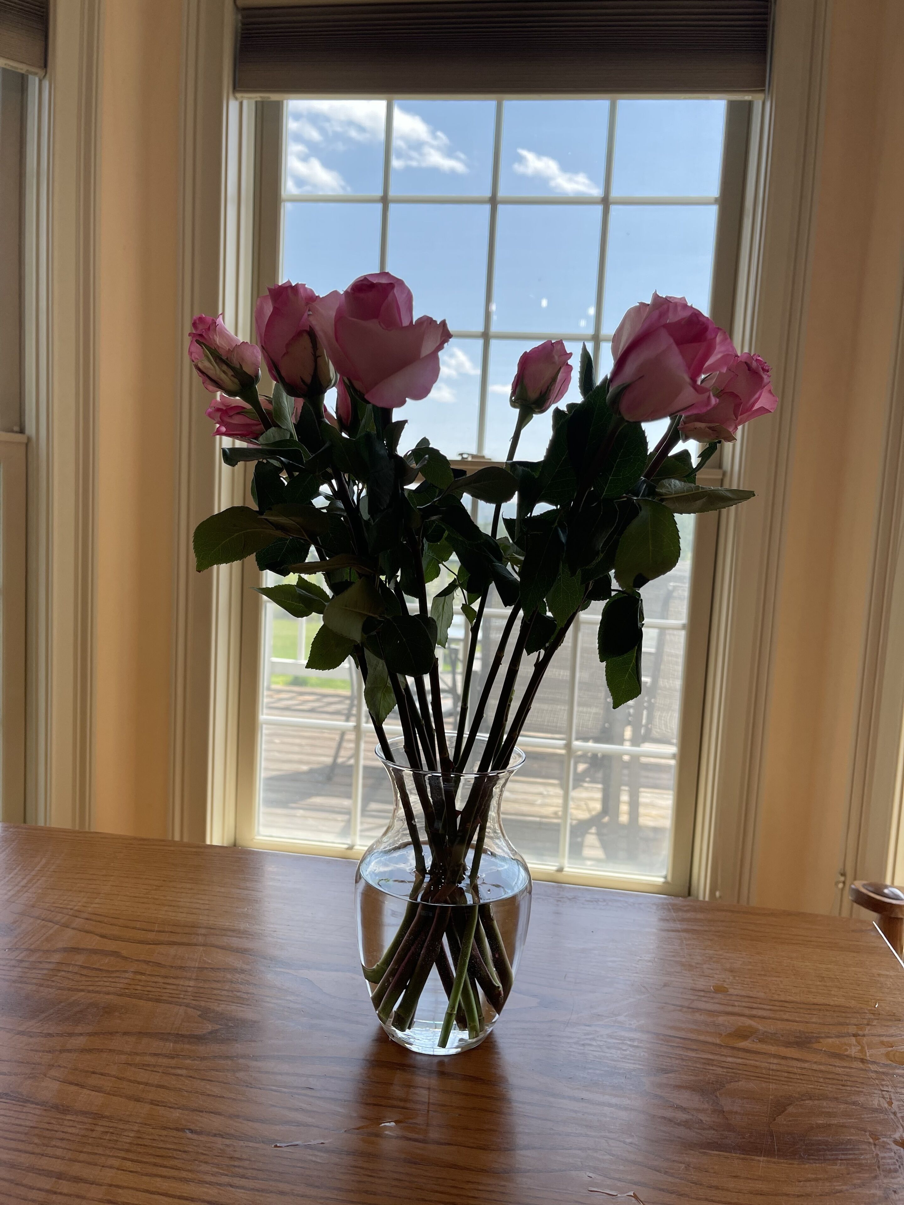 A clear vase with pink roses on a wooden table by a window.