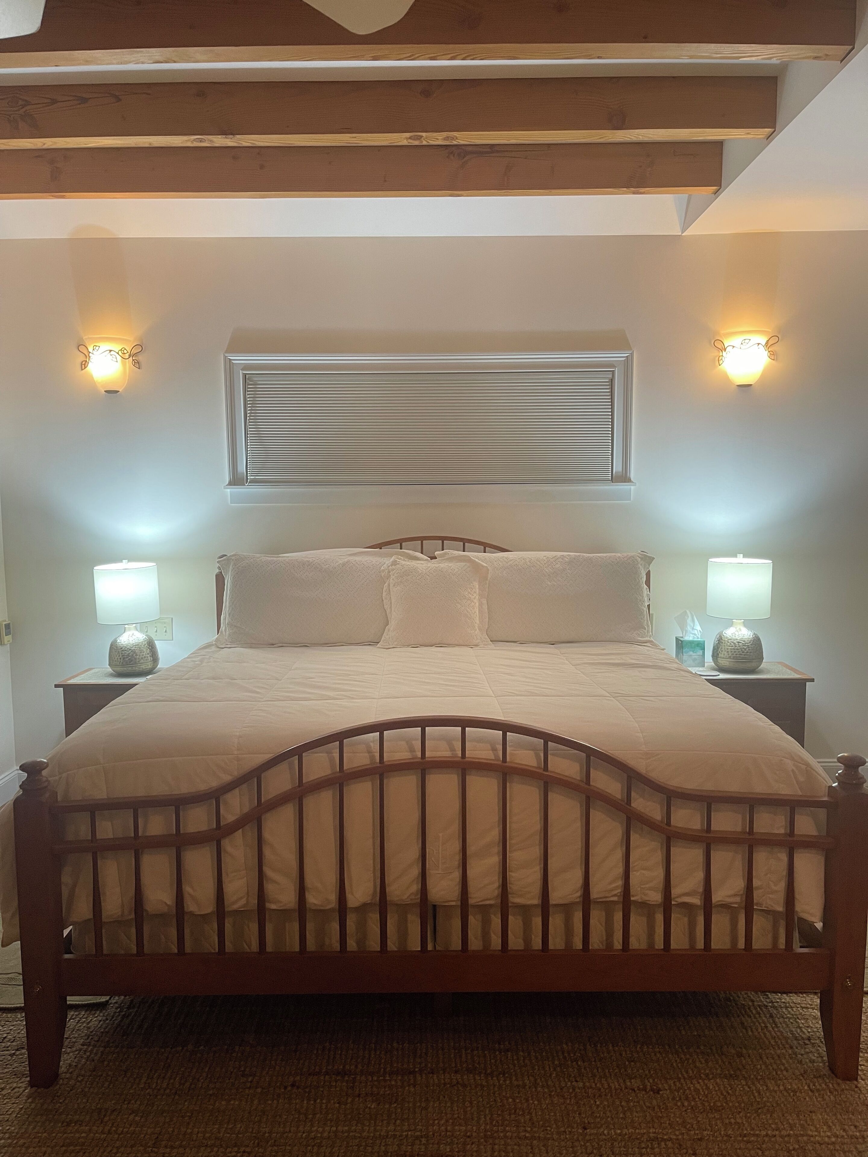 Un dormitorio con estructura de cama de madera, dos lámparas de noche, una ventana con persianas y un aplique de pared.