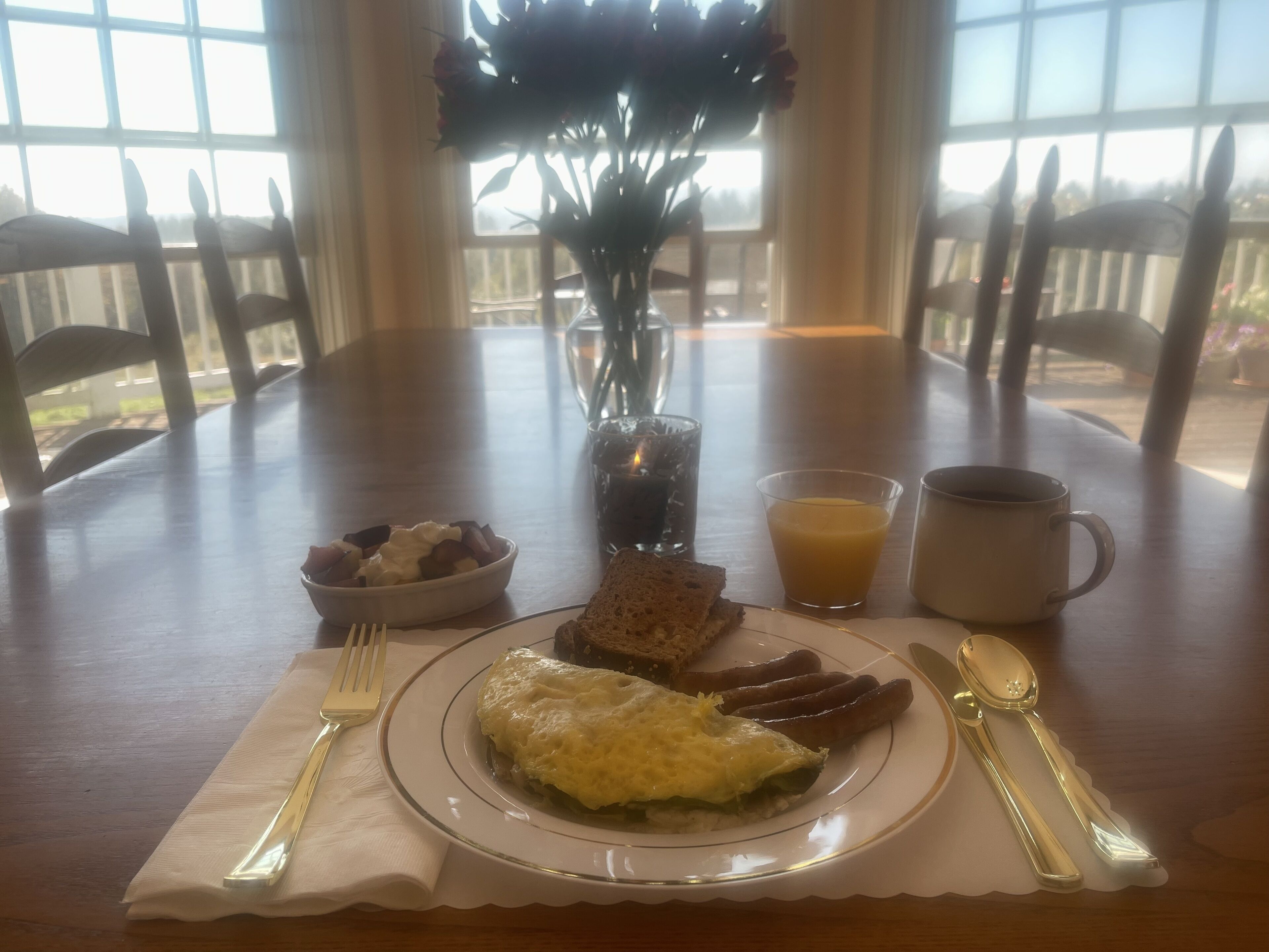 Una mesa de desayuno con un plato de huevos, salchichas, tostadas y un bol de fruta. Hay un vaso de zumo de naranja y una taza de café. Un jarrón con flores está sobre la mesa.
