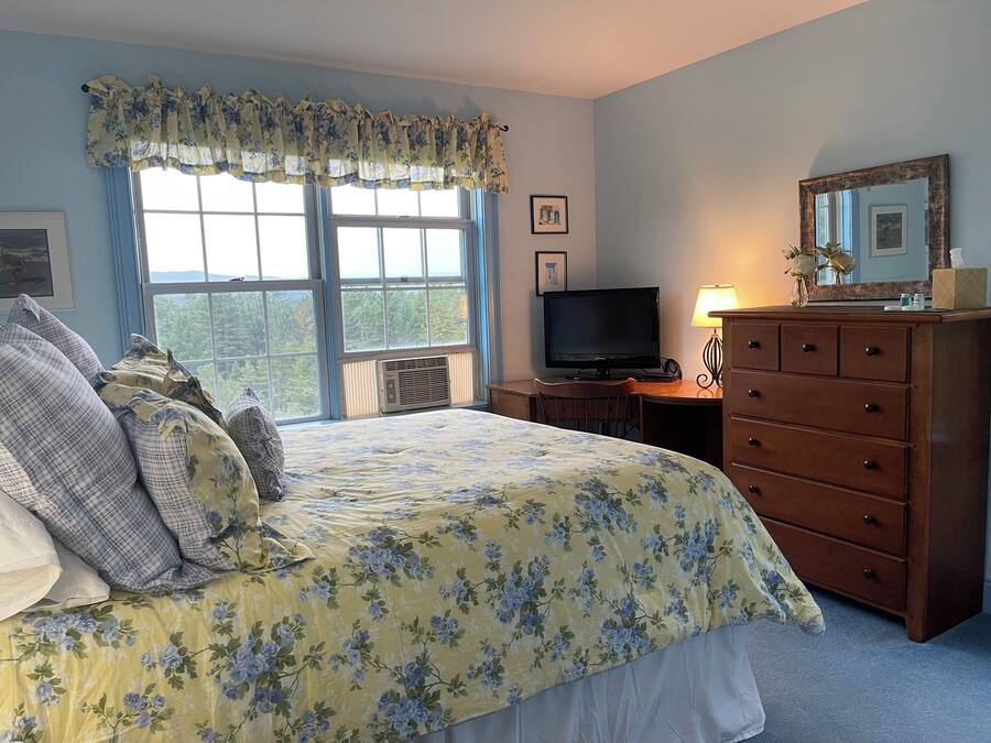 Un dormitorio con cama, televisor, cómoda y ventana con vista a árboles.