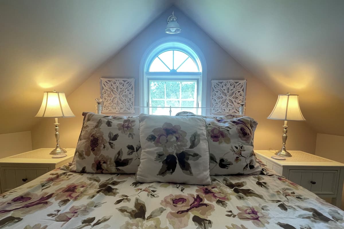 A bedroom with a bed, floral bedding, two bedside lamps, decorative wall panels, and a window with a view of greenery.