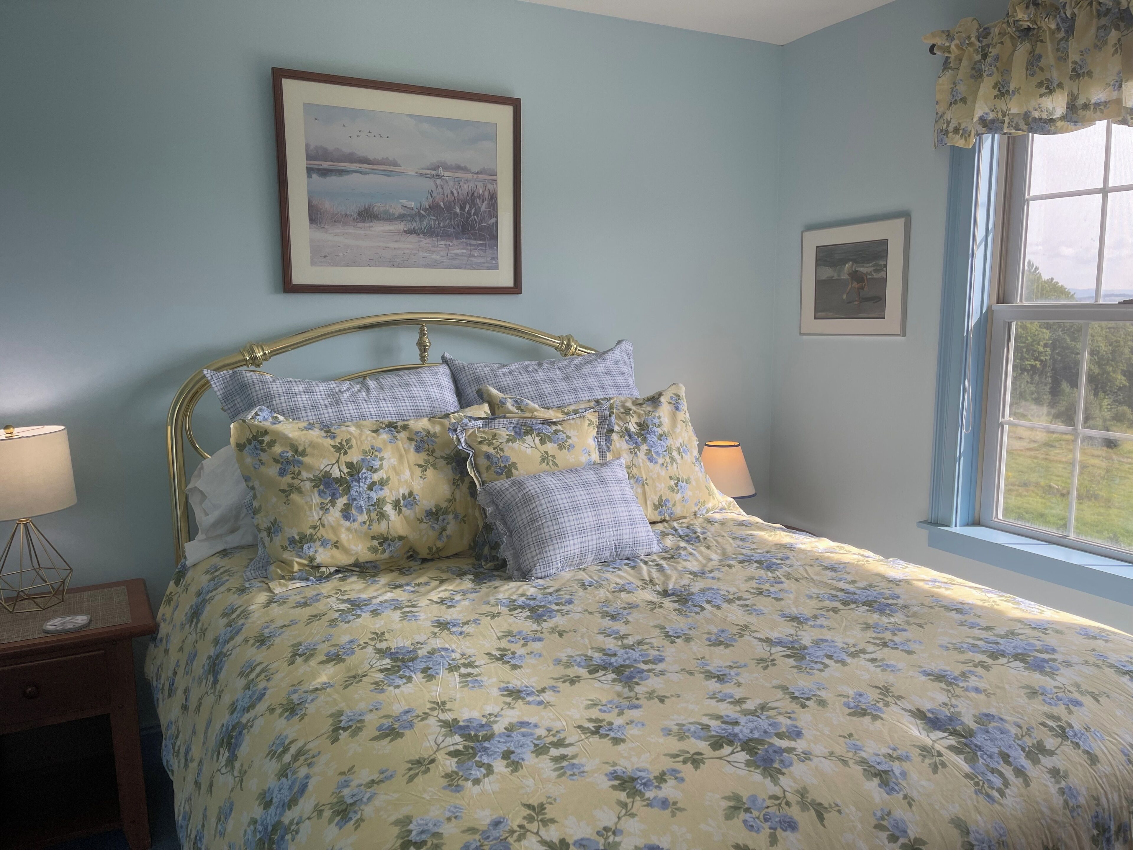 A bedroom with a floral bedspread, a metal-framed bed, two bedside lamps, and a window with a view of greenery.