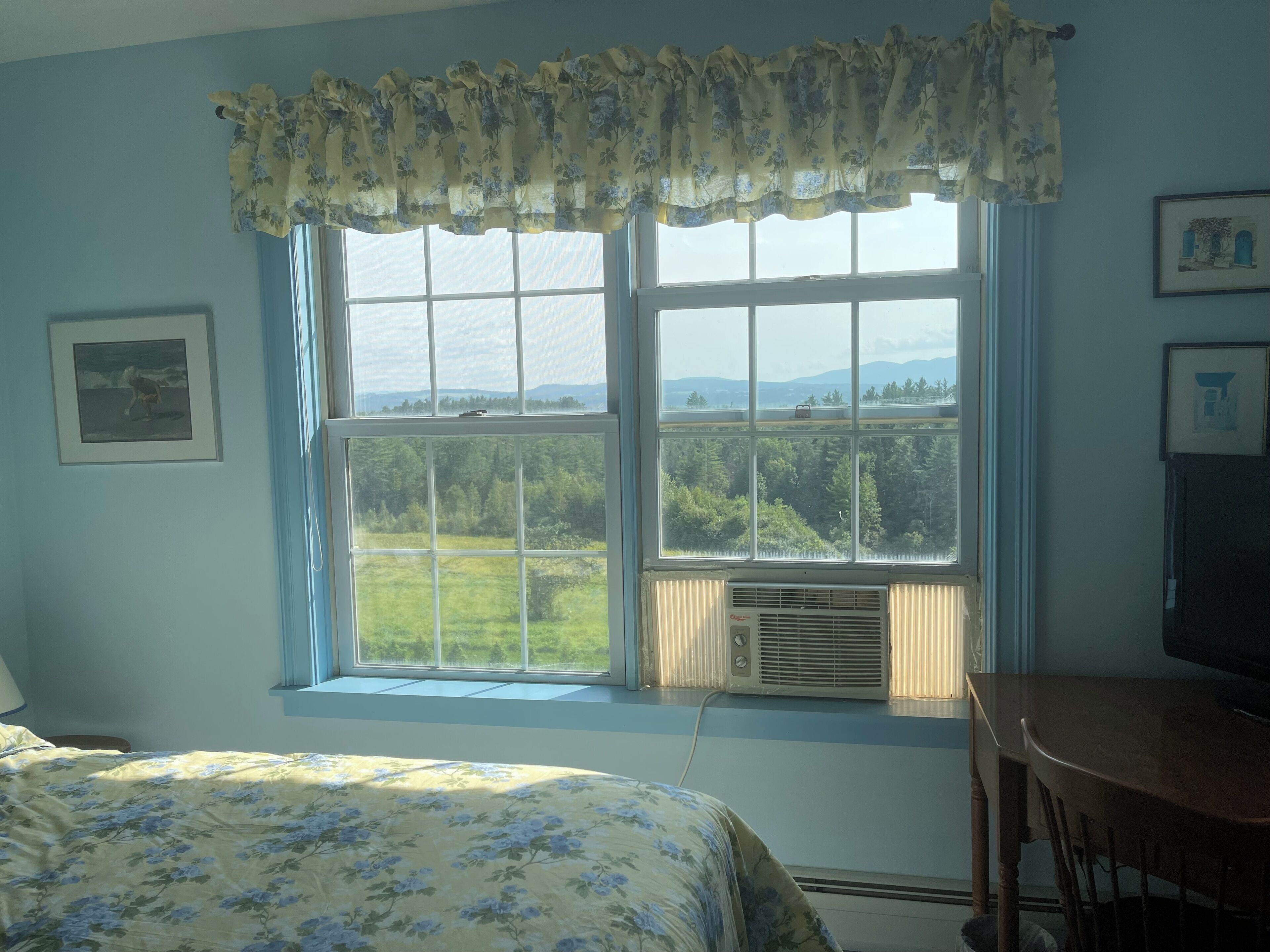 Un dormitorio con una ventana grande, una cama con ropa de cama floral, una silla y un cuadro en la pared.