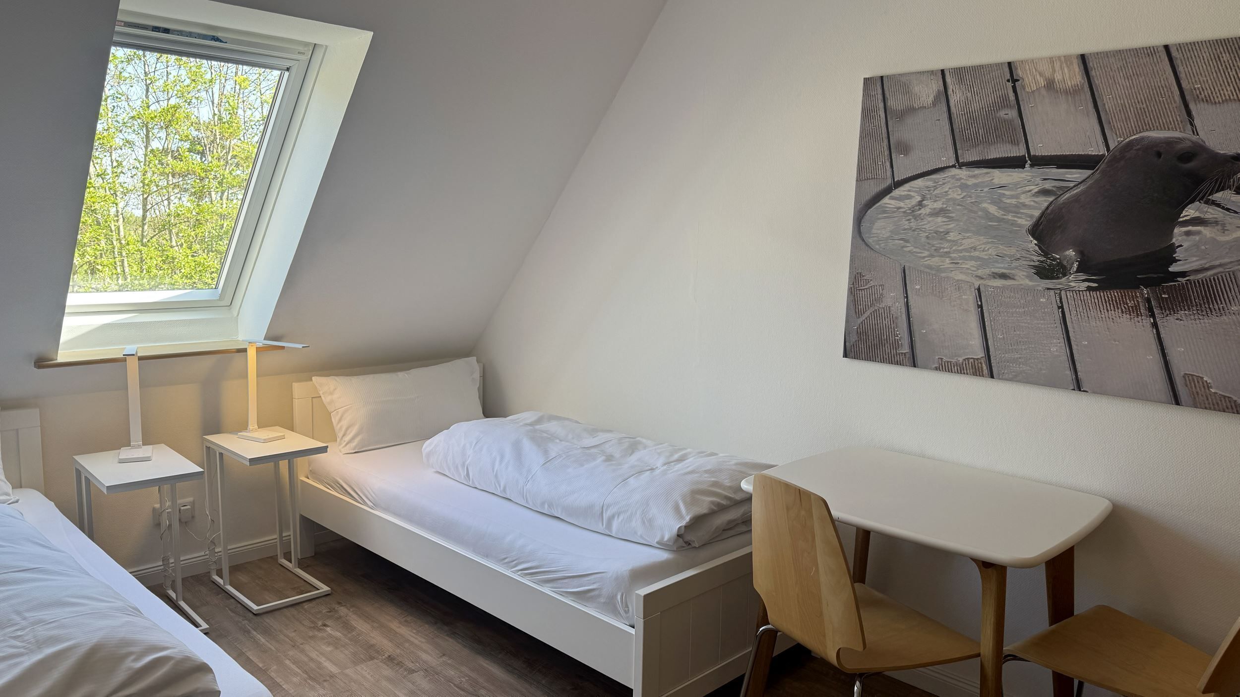 A small, modern room with a bed, a desk, and a chair. There is a window providing natural light and a painting on the wall.