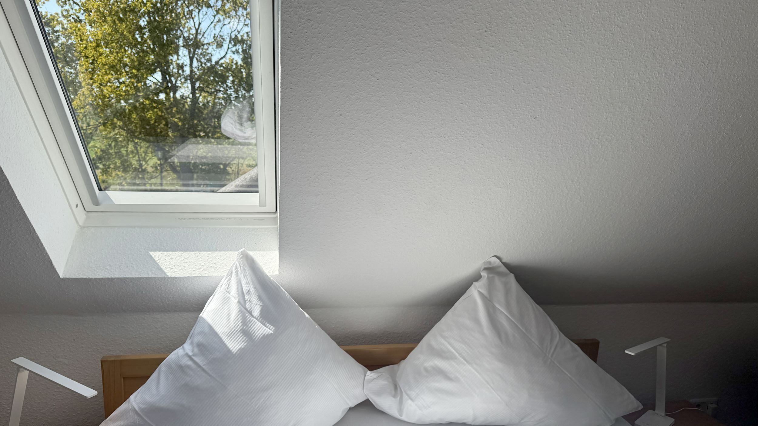 A room with a skylight, white walls, and two white pillows on a wooden bed.