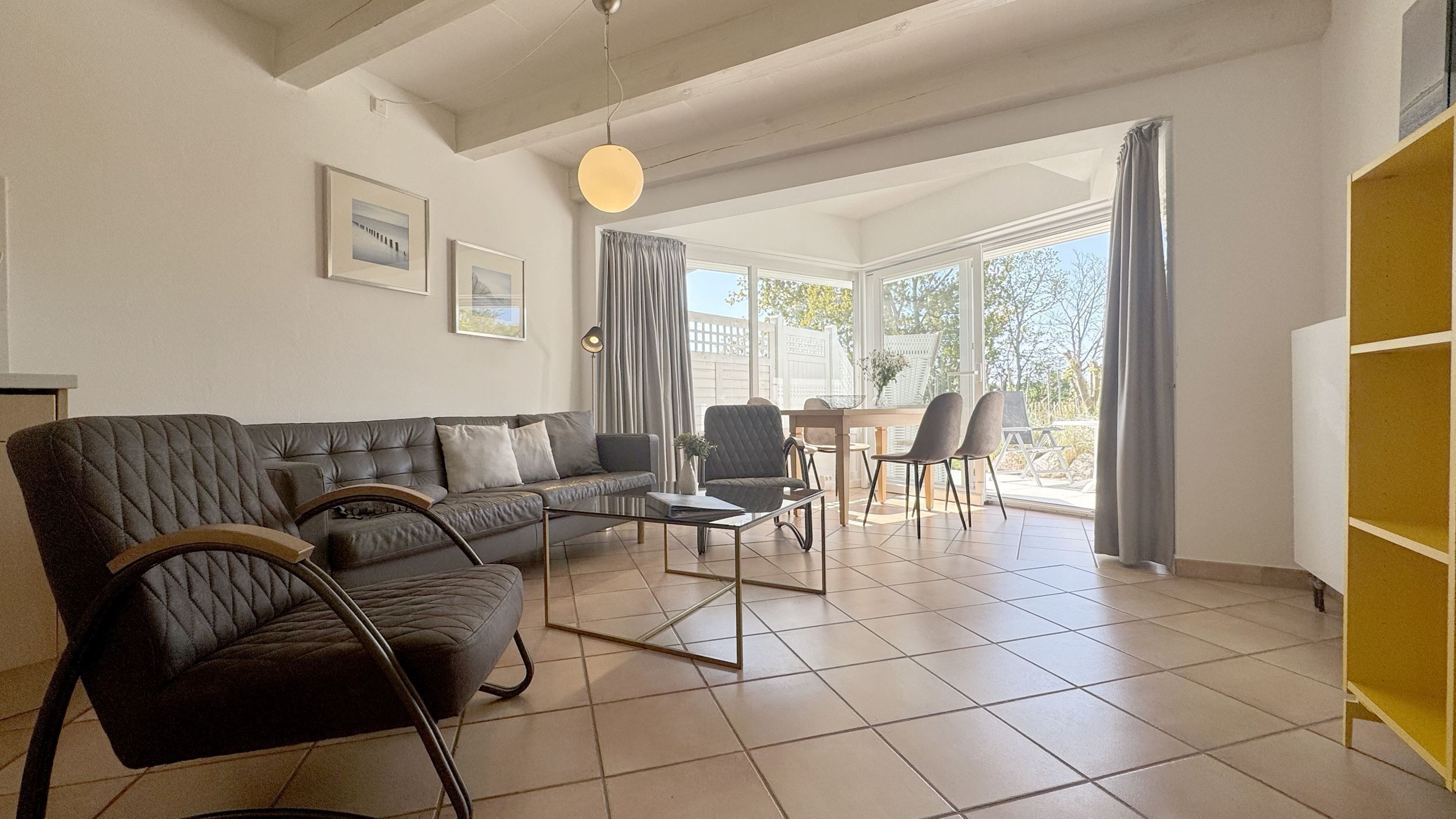 Un vaste salon comprenant un canapé d’angle, une table basse et un coin repas avec des chaises. De grandes fenêtres sont agrémentées de rideaux, et le salon est équipé d’un ventilateur de plafond et de tableaux encadrés qui ornent les murs.