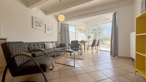 Un vaste salon comprenant un canapé d’angle, une table basse et un coin repas avec des chaises. De grandes fenêtres sont agrémentées de rideaux, et le salon est équipé d’un ventilateur de plafond et de tableaux encadrés qui ornent les murs.