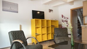 Un salon avec une bibliothèque jaune, deux fauteuils rembourrés, une table basse en verre et un téléviseur à écran plat fixé au mur.