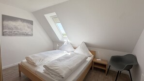 Une chambre avec un plafond en pente, un puits de lumière, un lit avec du linge de maison blanc, une table de chevet, une chaise et un tableau accroché au mur.