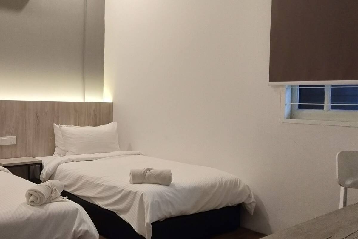 A hotel room with two beds, a small table, a chair, and a window with blinds.