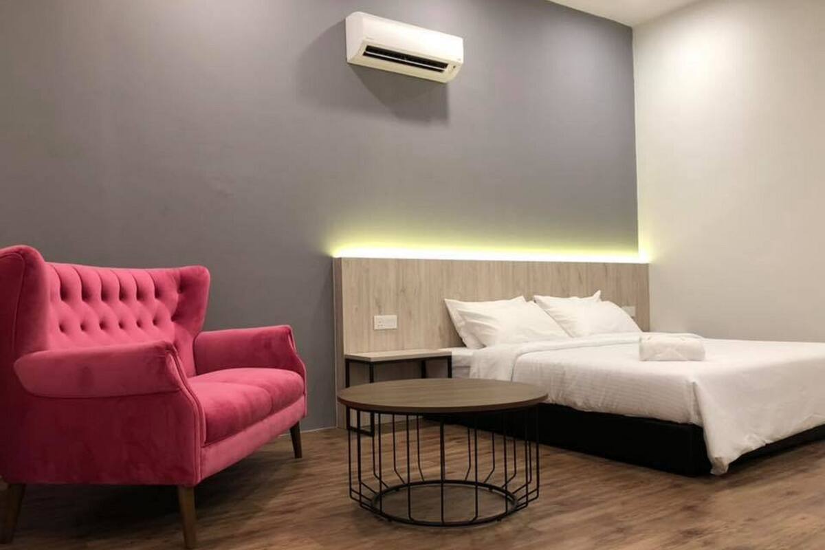 A modern hotel room with a tufted pink chair, a round wooden table, a bed with white linens, and a wall-mounted air conditioner.