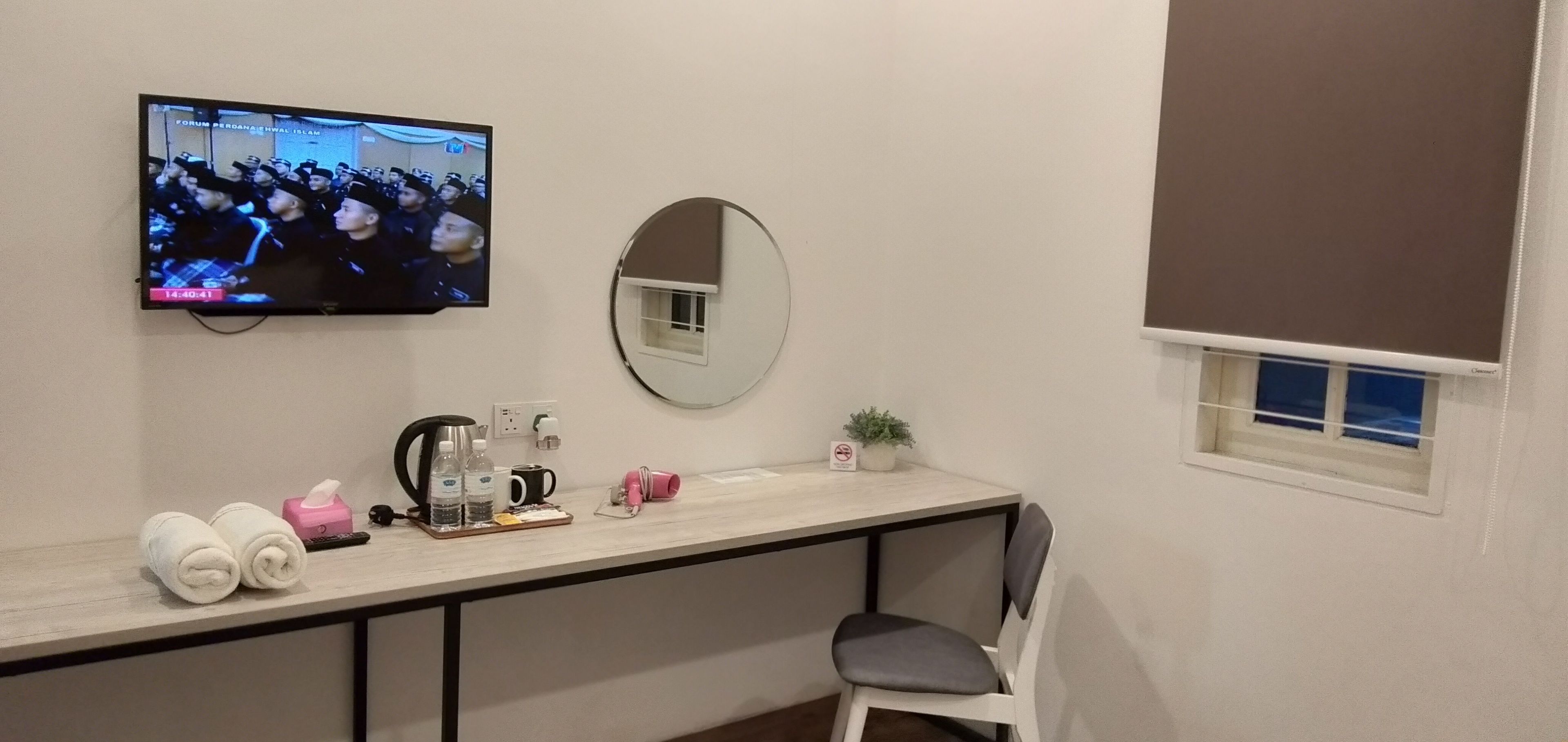 A hotel room with a TV mounted on the wall, a round mirror, a small window, a desk with toiletries, and a chair.