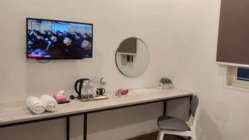 A hotel room with a TV mounted on the wall, a round mirror, a small window, a desk with toiletries, and a chair.