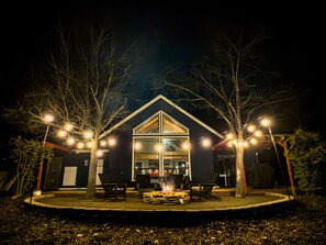 Une maison moderne avec un foyer extérieur dans le jardin, entourée d’arbres et illuminée de guirlandes lumineuses.