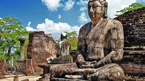 Eine große Buddha-Statue aus Stein in einer historischen Stätte, mit alten Backgebäuden und Bäumen im Hintergrund.