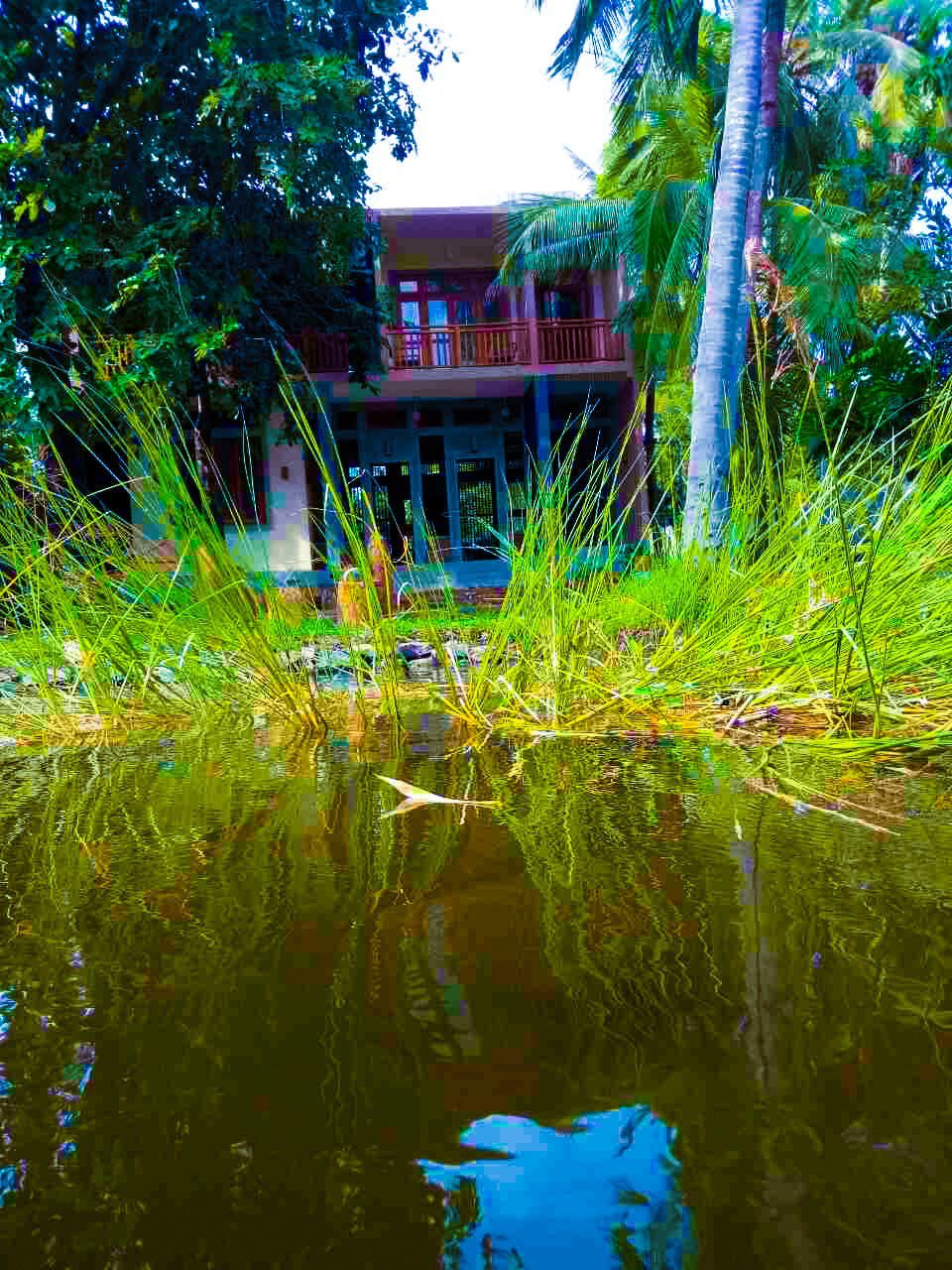 A building with a balcony, surrounded by lush greenery and a body of water reflecting the structure.
