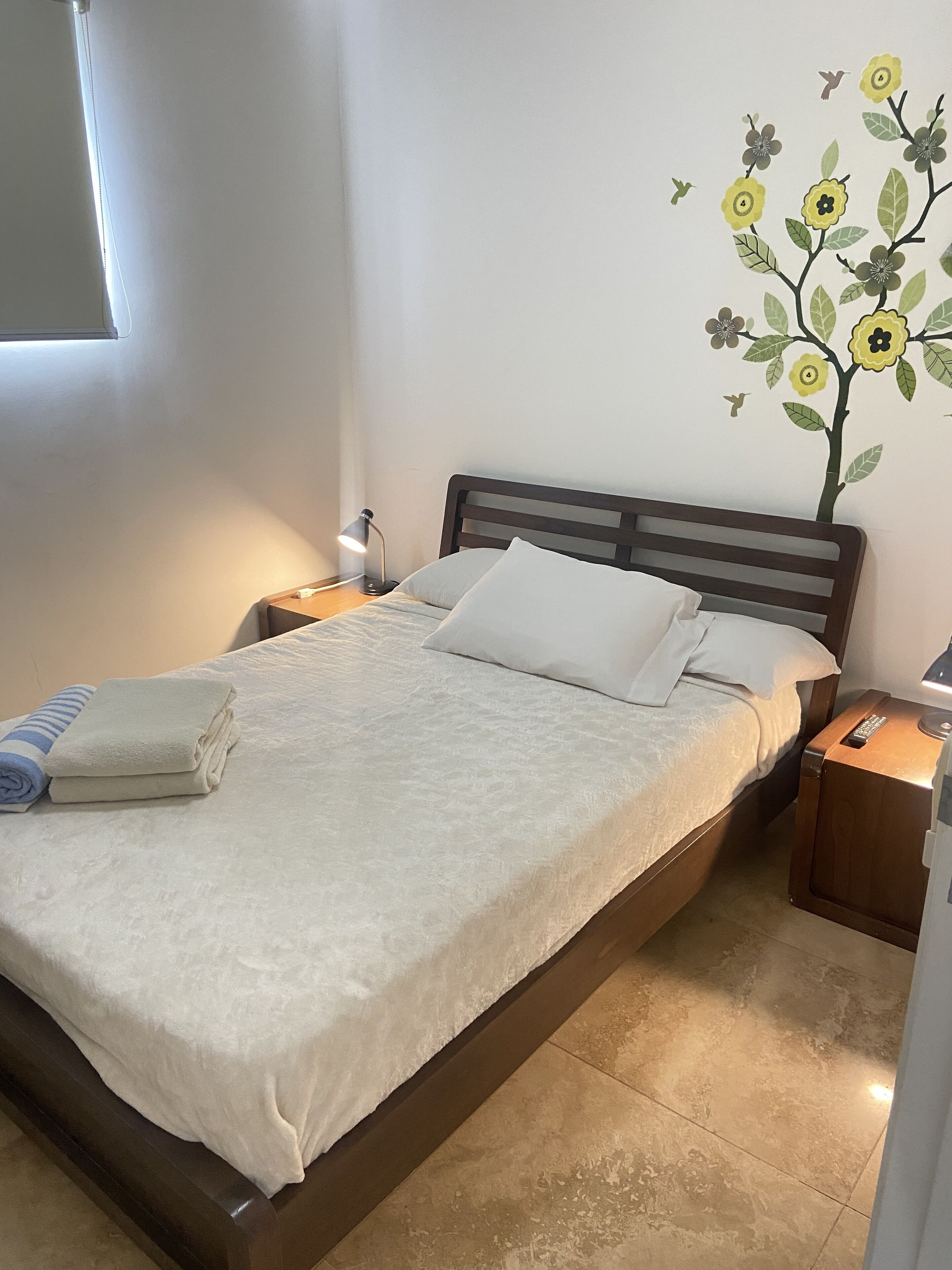 A neatly made bed with a white duvet, a folded towel, and a remote control on a wooden nightstand beside it.
