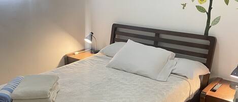 A neatly made bed with a white duvet, a folded towel, and a remote control on a wooden nightstand beside it.