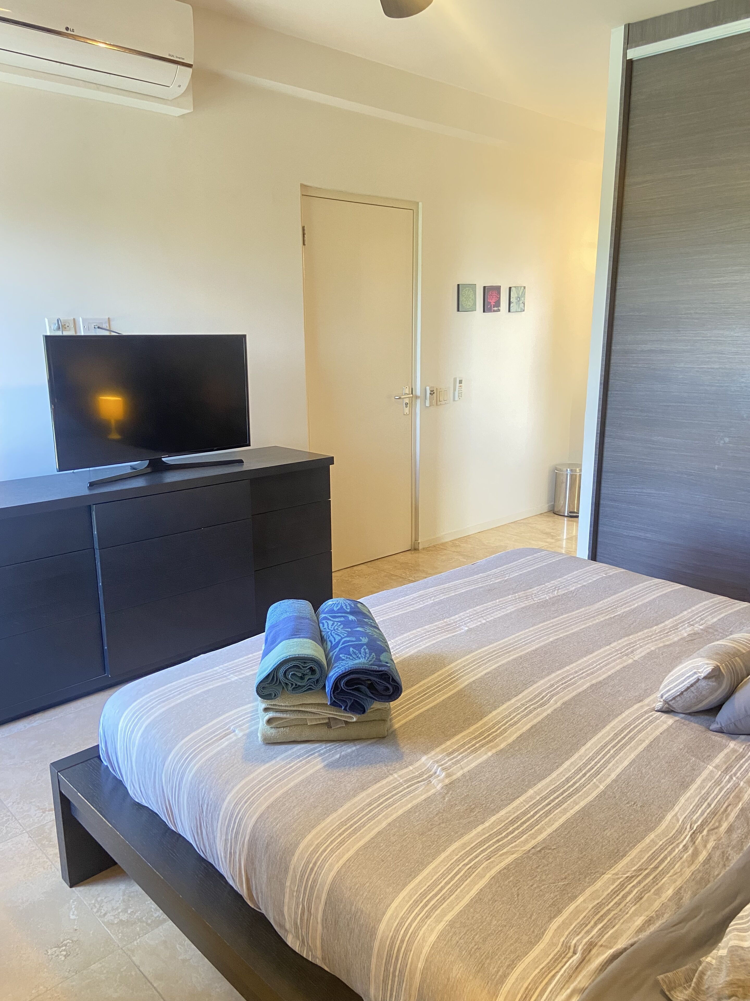 Une chambre à coucher avec un lit, un téléviseur posé sur une commode, une porte et un mur orné de trois petits tableaux.