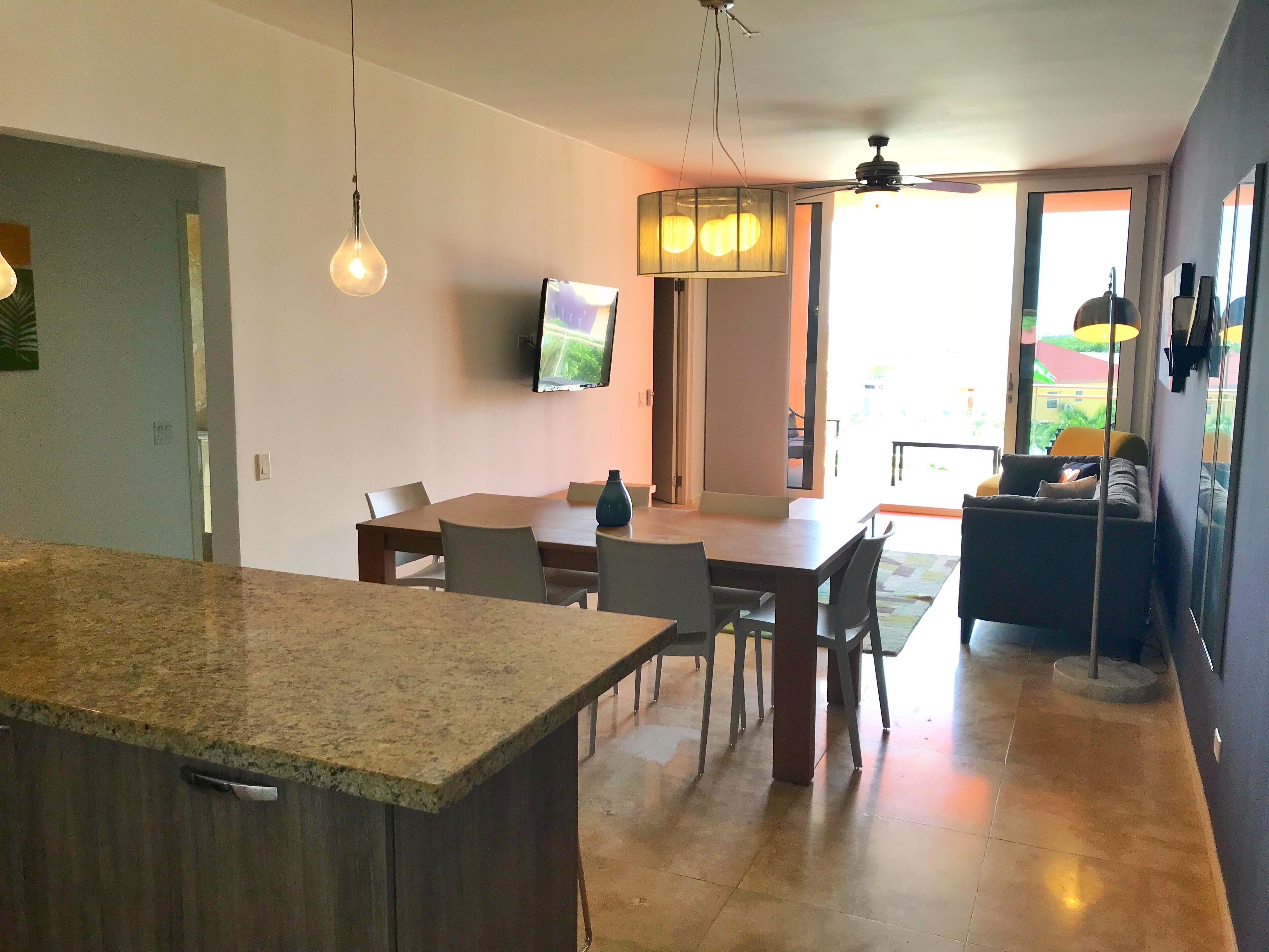 A modern kitchen with granite countertops, a dining area with a table and chairs, a flat-screen TV mounted on the wall, and a ceiling fan.