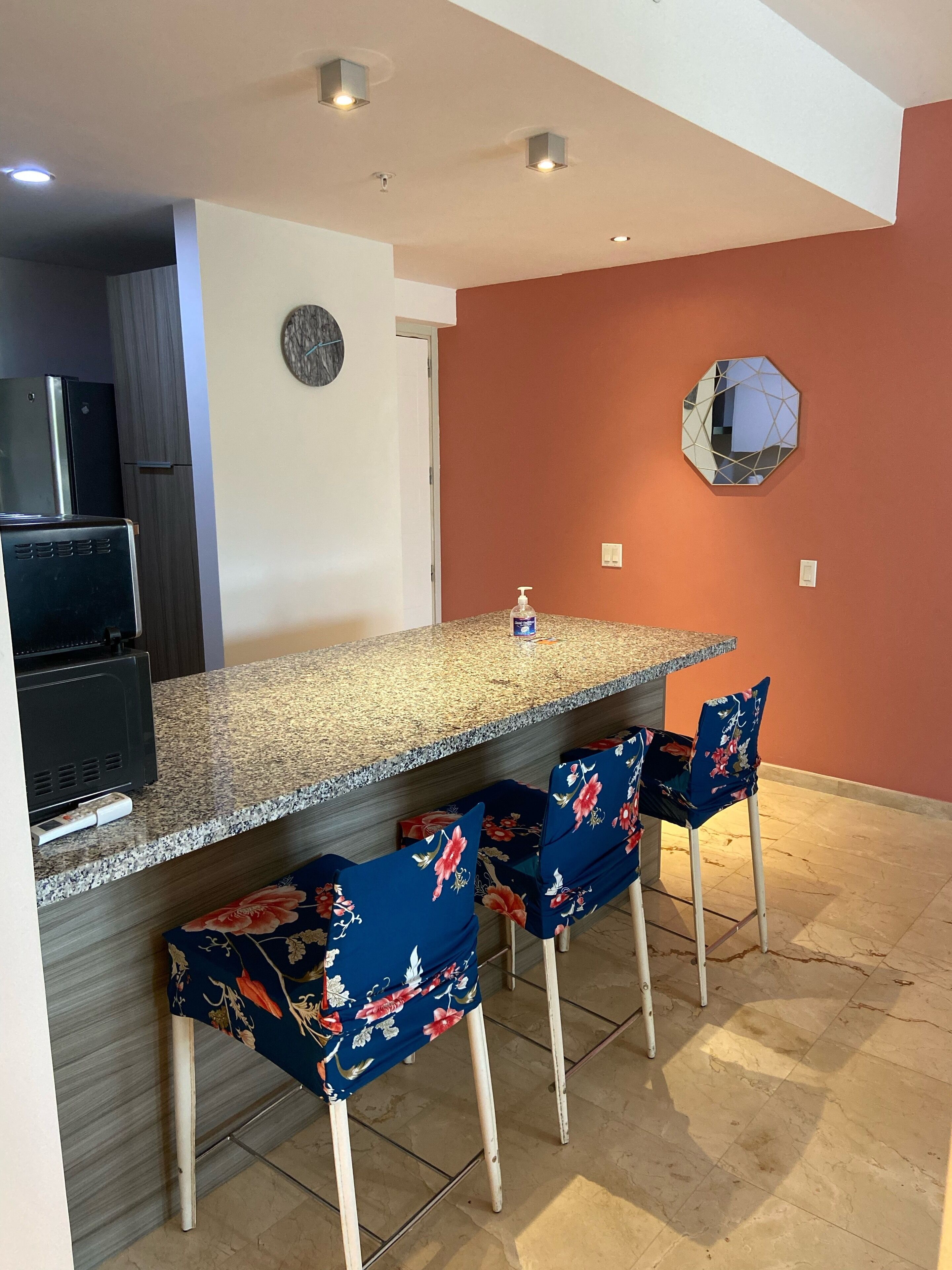 A kitchen with a granite countertop, a built-in oven, and a refrigerator. There are three bar stools with floral upholstery.