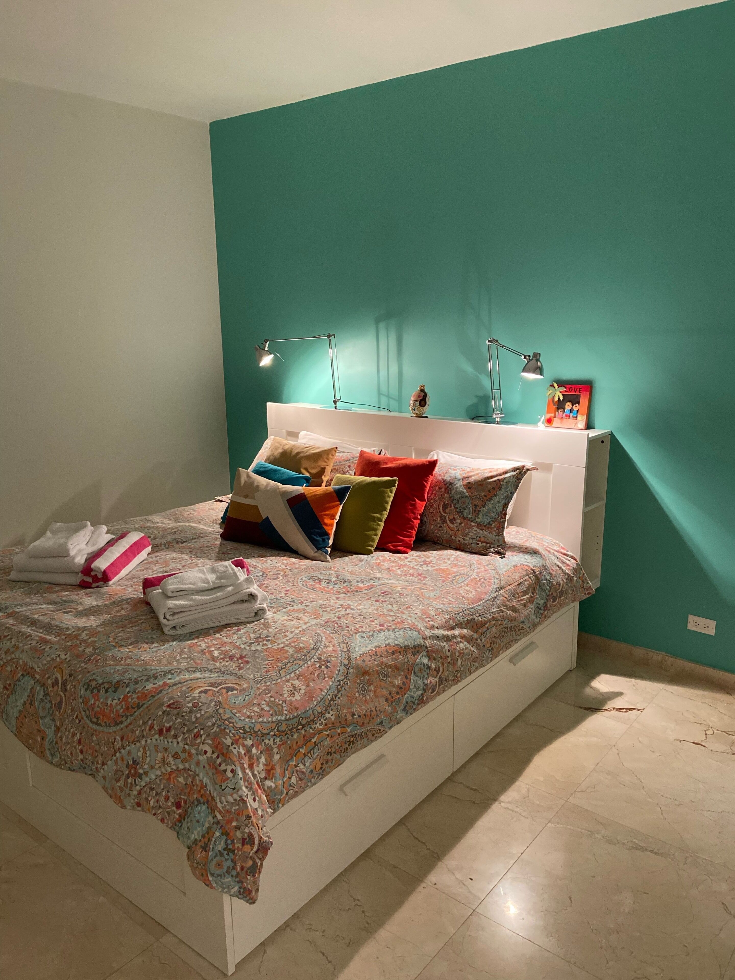 Une chambre à coucher avec un lit, des oreillers, une table de chevet, une lampe et un mur de couleur turquoise.