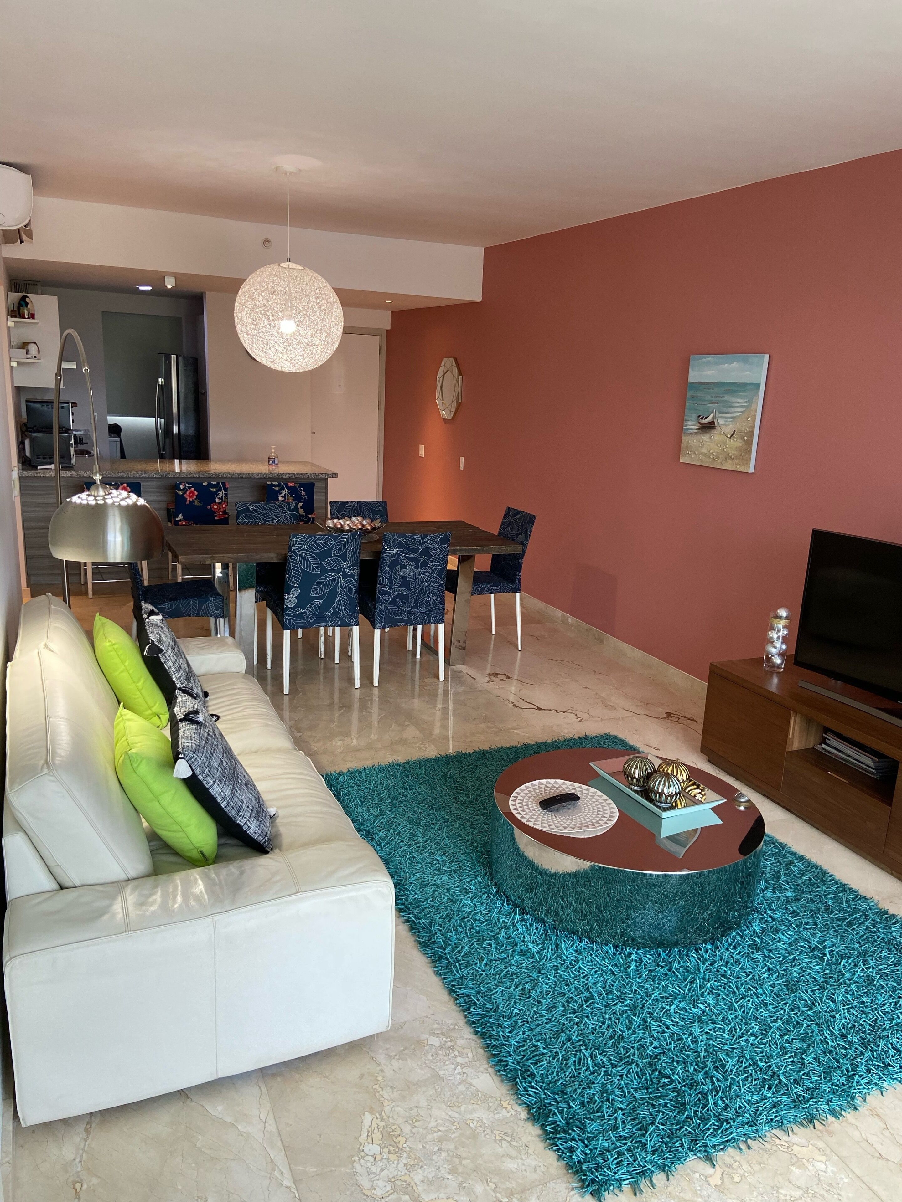 A modern living room with a white sofa, teal area rug, and a dining area with blue chairs.