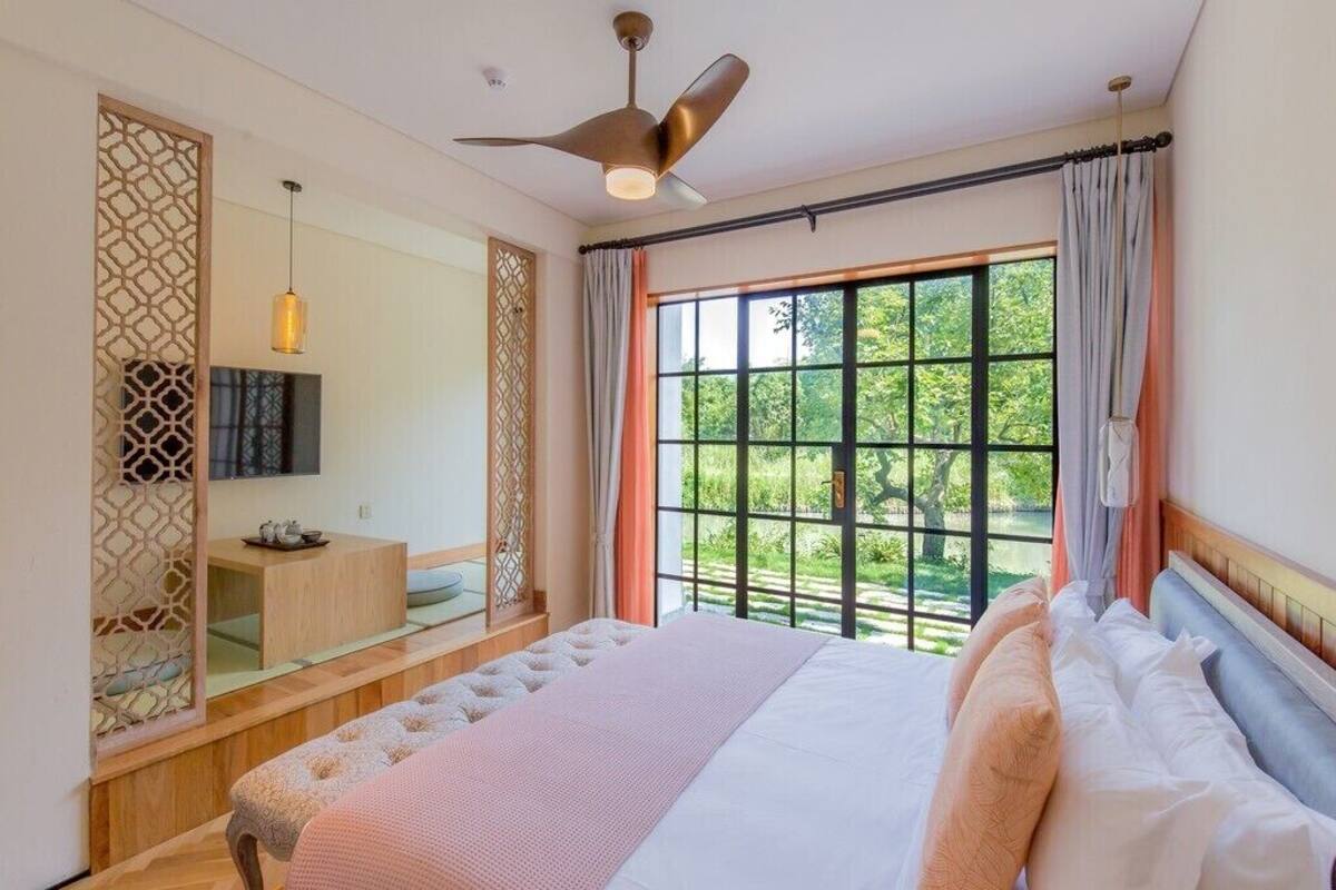 Quarto com cama, ventilador de teto, televisão, janela com cortinas e vista para o verde lá fora.