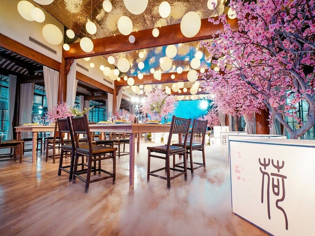 A restaurant with wooden tables and chairs, a ceiling with hanging lights, and a large tree with pink blossoms.
