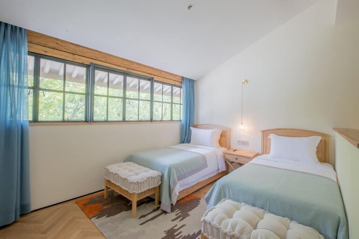 Quarto com duas camas, janela com cortinas, teto de madeira e mesa de cabeceira com abajur.