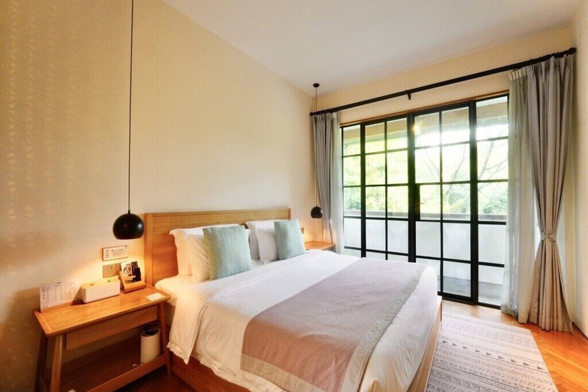 Quarto com cama grande, mesa de cabeceira com abajur, janela com cortinas e vista para o verde lá fora.