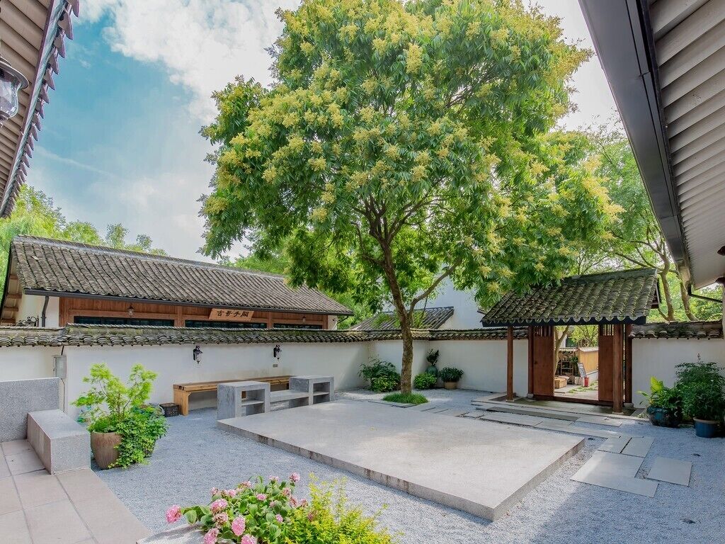 Un patio japonés tradicional con un gran árbol, bancos de piedra y un edificio de madera con tejado de tejas.