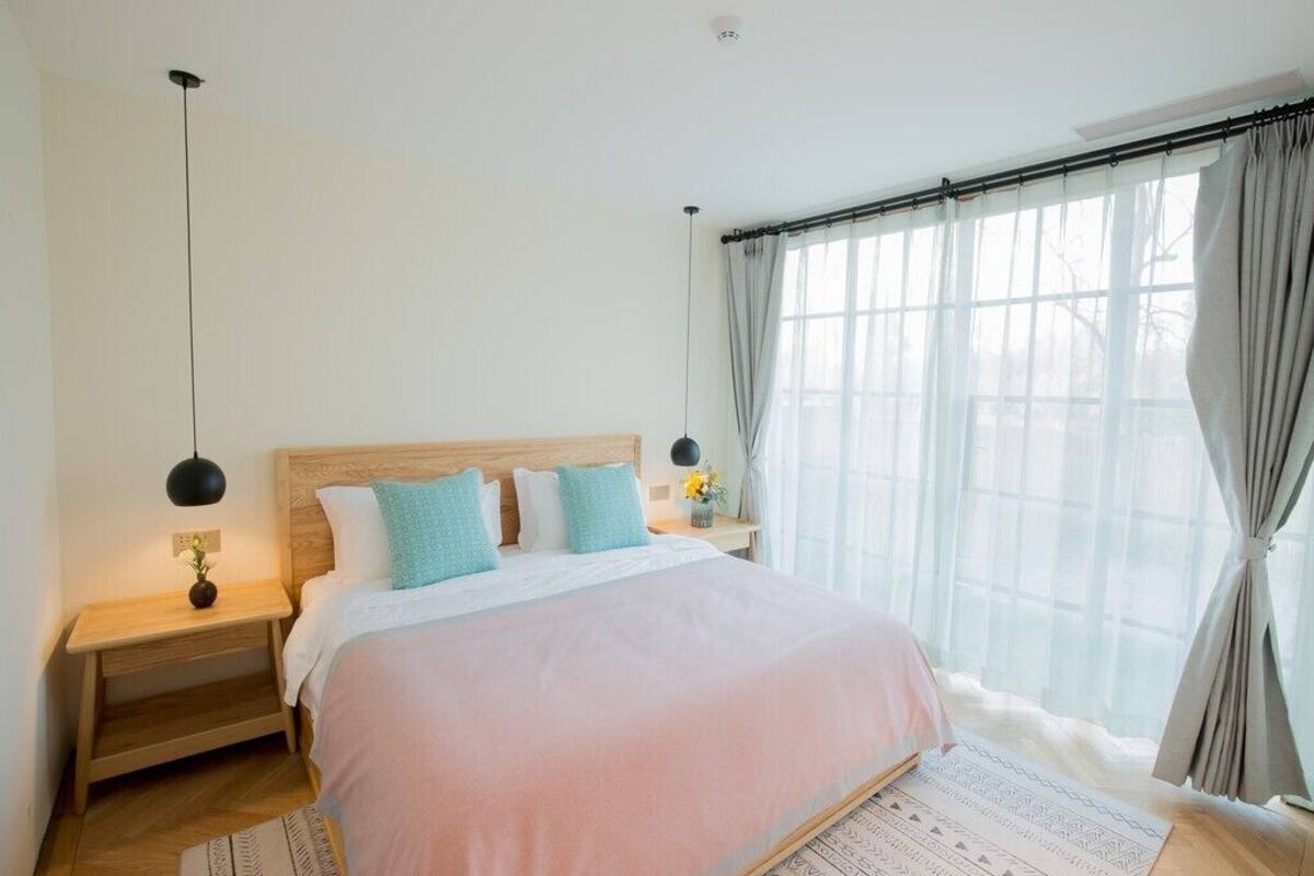 Quarto com cama grande, cabeceira de madeira, mesas de cabeceira, um criado-mudo, janela com cortinas e luminária pendente.