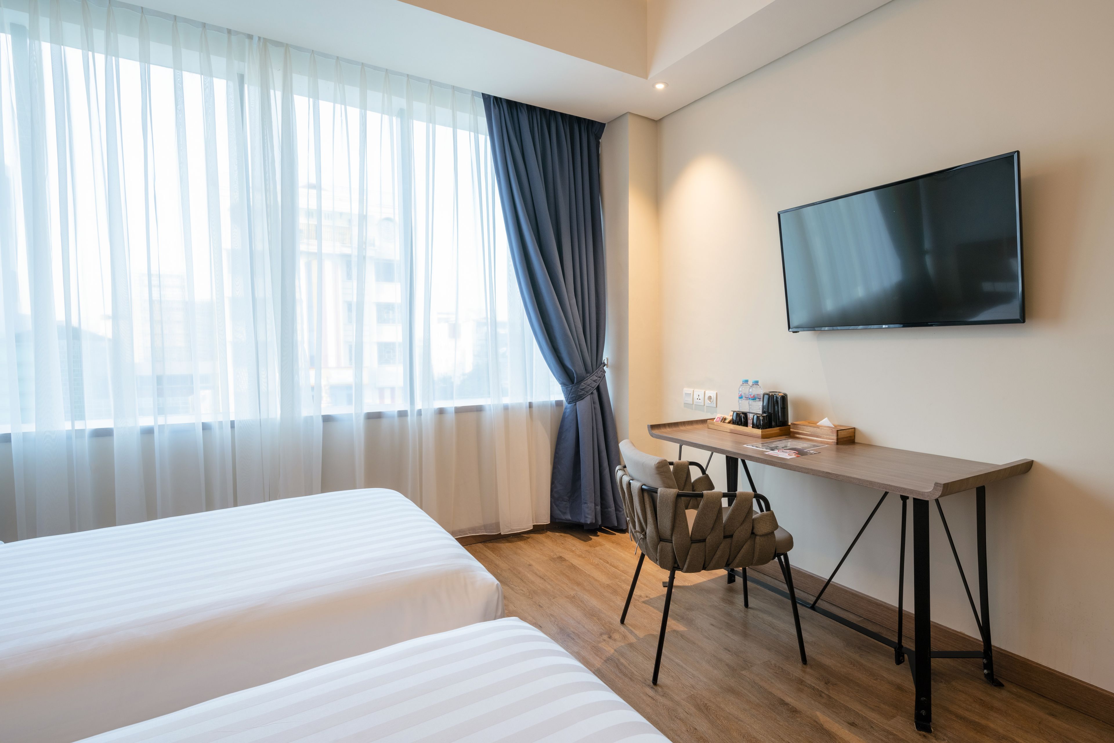 A hotel room with two beds, a desk, a chair, a TV, and a window with sheer curtains.