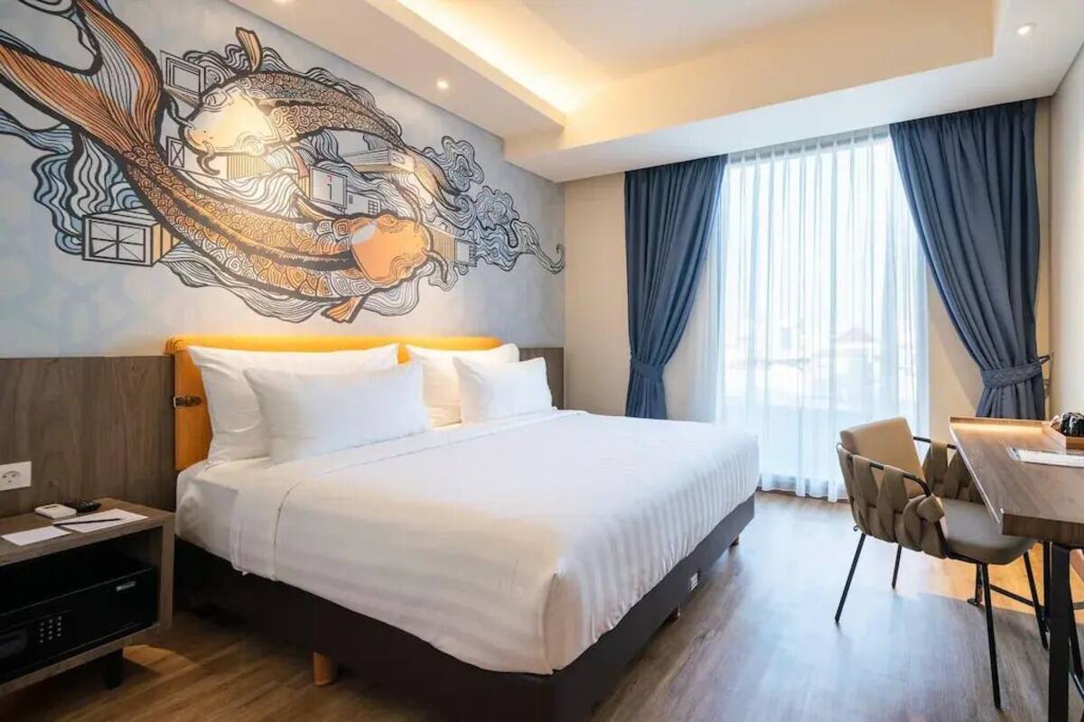 A hotel room with a large bed, a desk with chairs, a wall mural, and a window with curtains.