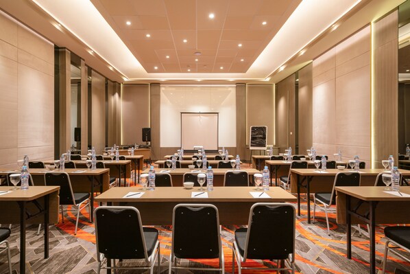 Un salón de conferencias con varias mesas y sillas, equipado con botellas de agua y vasos, y una pantalla para presentaciones al frente.