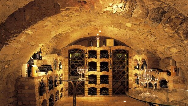 Una bodega subterránea con arcos de piedra, estanterías para vino y una mesa de cristal.