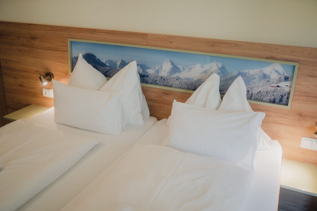 A hotel room with a wooden headboard, two white beds with pillows, a nightstand, and a wall-mounted painting of a mountain landscape.