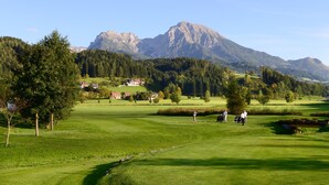 Un campo de golf con montañas al fondo, algunos árboles dispersos y un pequeño edificio a lo lejos.