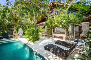 Un espace piscine doté de chaises longues, d’une petite table et d’un fauteuil suspendu, le tout entouré d’une végétation luxuriante et d’un bâtiment dont le toit est recouvert de tuiles.