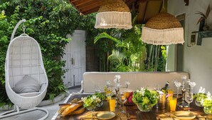An outdoor dining area with a wicker chair, a cushioned bench, and a table set for a meal. - Luxury Enclosed Villa CHARM in Seminyak - 3 min walk to the beach (Seminyak)