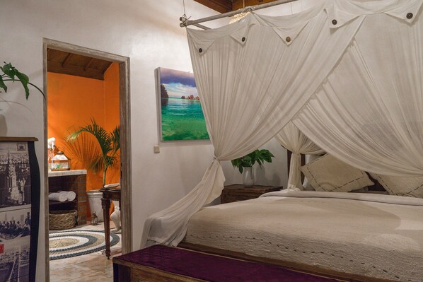 Une chambre à coucher avec un lit à baldaquin, un tableau représentant une scène de plage, une plante et un téléphone d’époque.