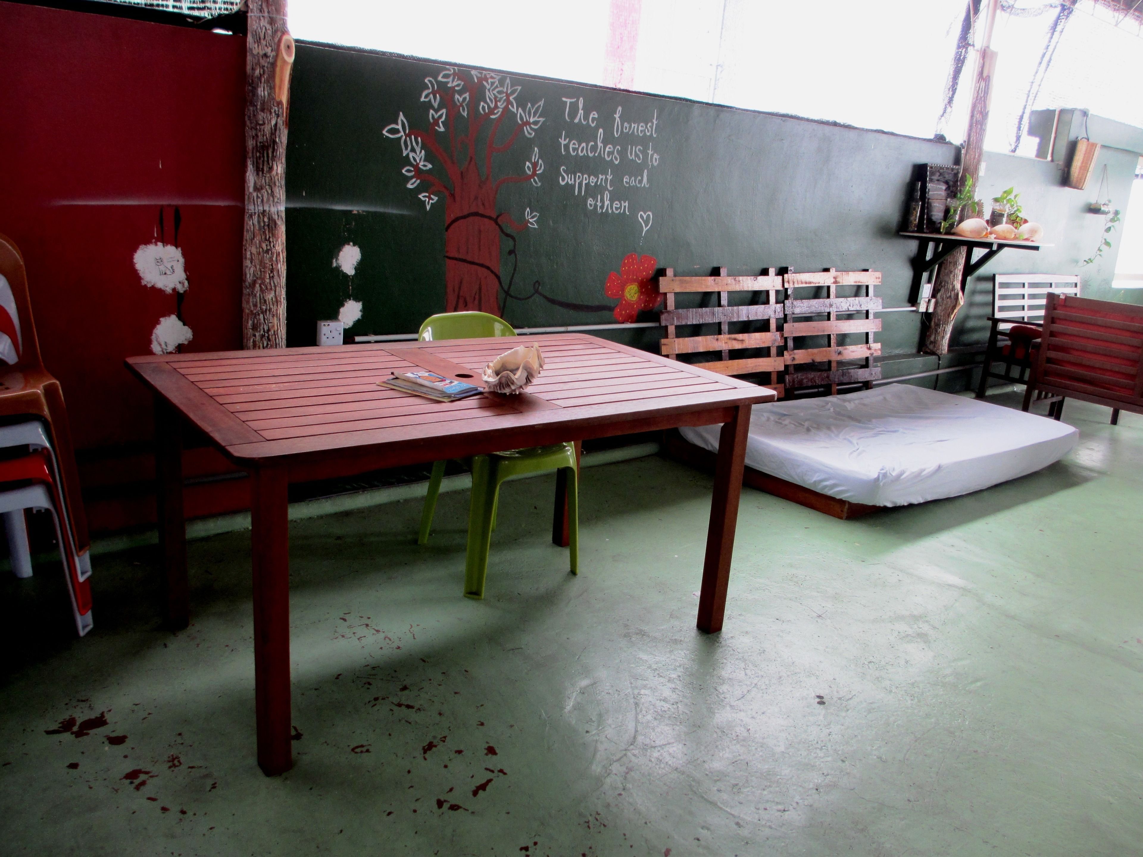 A room with a wooden table, a green chair, a mattress, and a chalkboard with a tree drawing and text.