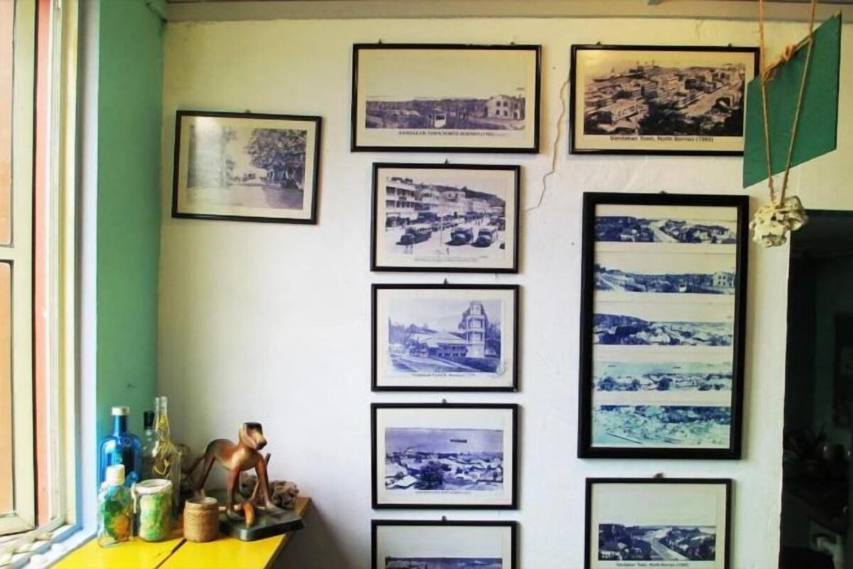 Un mur orné de plusieurs photographies en noir et blanc encadrées, une petite table avec des bouteilles et une figurine de singe, et une fenêtre avec un cadre vert.