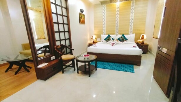 A hotel room with a bed, bedside tables, a small round table, a chair, and a large window. - Hotel Ozas Grand (Varanasi)