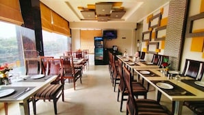 A restaurant with long tables and chairs, a Pepsi refrigerator, and a TV on the wall. - Hotel Ozas Grand (Varanasi)