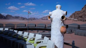 A desert landscape with a white structure resembling a fire hydrant, outdoor seating with green cushions, and a flag in the background. - Desert Sand Camp (Wadi Rum)